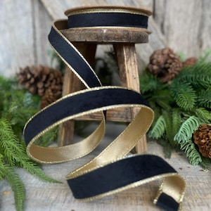 May include: Black velvet ribbon with gold trim. The ribbon is rolled up on a wooden stool and is surrounded by green pine branches.