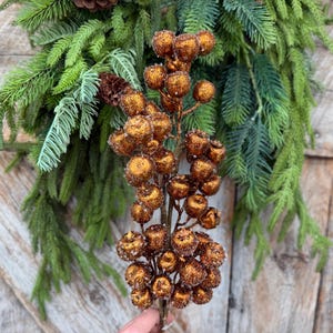 May include: A decorative floral pick featuring a cluster of round, glittery copper-colored berries on a central stem, positioned in front of a lush arrangement of green pine needles and small brown pinecones.