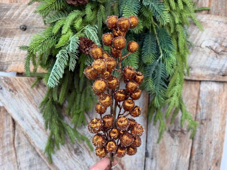 May include: A decorative floral pick featuring a cluster of round, glittery copper-colored berries on a central stem, positioned in front of a lush arrangement of green pine needles and small brown pinecones.