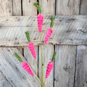 May include: A cluster of pink, faux carrots with green stems and leaves. The carrots are arranged on thin green stems against a weathered wooden background. The carrots are a vibrant pink color and have a textured surface.