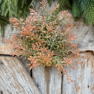 May include: A close-up of a decorative floral arrangement featuring a cluster of orange and green leaves. The arrangement is set against a rustic wooden background, with evergreen branches and pinecones in the background.