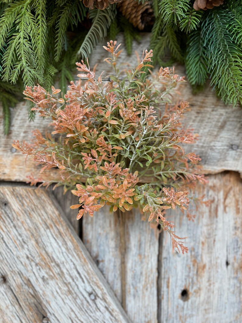 May include: A close-up of a decorative floral arrangement featuring a cluster of orange and green leaves. The arrangement is set against a rustic wooden background, with evergreen branches and pinecones in the background.