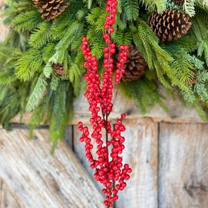 May include: A sprig of bright red berries with a brown stem, set against a backdrop of a Christmas wreath with pinecones and evergreen branches. The berries are clustered closely together, creating a vibrant contrast with the green foliage.