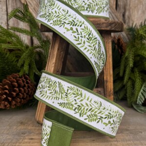 May include: A spool of white fabric ribbon featuring a repeating green watercolor fern leaf pattern. The ribbon has finished green edges and is wound around a wooden spool, displaying a continuous botanical print of various green foliage.