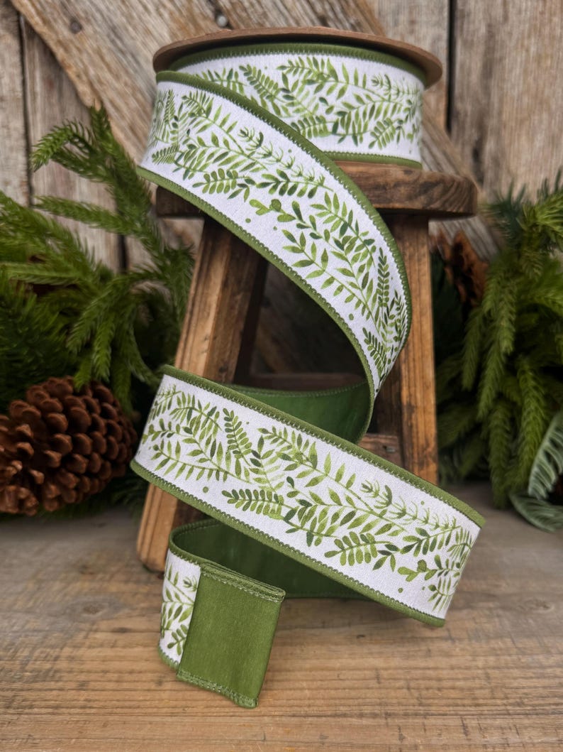 May include: A spool of white fabric ribbon featuring a repeating green watercolor fern leaf pattern. The ribbon has finished green edges and is wound around a wooden spool, displaying a continuous botanical print of various green foliage.