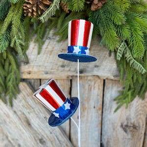 May include: Two red, white, and blue patriotic top hats with glitter on the blue brim. The hats are attached to white stems.