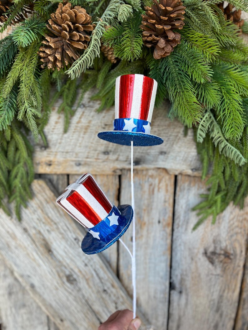 May include: Two red, white, and blue patriotic top hats with glitter on the blue brim. The hats are attached to white stems.