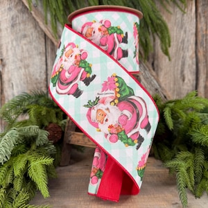 May include: A spool of wired ribbon featuring a light mint green and white buffalo check pattern. The ribbon is decorated with illustrations of Santa Claus wearing a pink suit and holding a green wreath, with the text Merry Christmas printed.