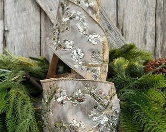 May include: A roll of decorative ribbon with a light gray fabric base. The ribbon is embellished with gold-colored sequins and embroidery in a floral pattern. The ribbon is displayed on a wooden stand, with evergreen branches and a pinecone in the background.