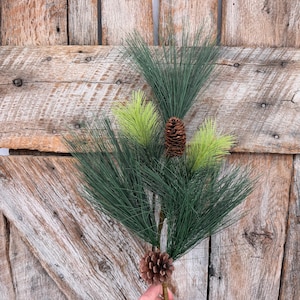 May include: A decorative artificial pine branch featuring long, thin dark green needles mixed with lighter lime green accents. The branch includes two brown pine cones, one positioned near the center and one at the base of the stem.