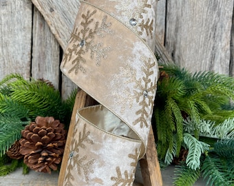 May include: A spool of beige velvet ribbon with gold glitter snowflake designs and crystal accents. The ribbon is displayed on a wooden stand, with pinecones and greenery in the background. This decorative ribbon is perfect for holiday crafts and gift wrapping.