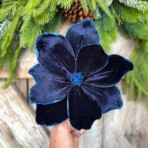 May include: A large, navy blue velvet flower with glittered edges. The flower is the focal point, set against a backdrop of evergreen branches and a pinecone, suggesting a holiday or seasonal theme.