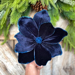 May include: A large, navy blue velvet flower with glittered edges. The flower is the focal point, set against a backdrop of evergreen branches and a pinecone, suggesting a holiday or seasonal theme.