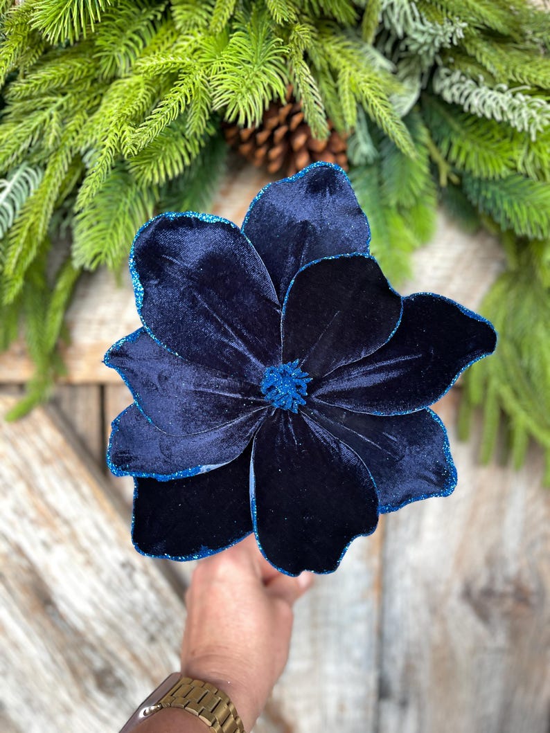 May include: A large, navy blue velvet flower with glittered edges. The flower is the focal point, set against a backdrop of evergreen branches and a pinecone, suggesting a holiday or seasonal theme.