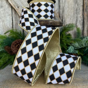 May include: Decorative ribbon with a black and white harlequin pattern and gold trim. A roll of the same ribbon sits in the background. The ribbon is displayed on a wooden surface with greenery.