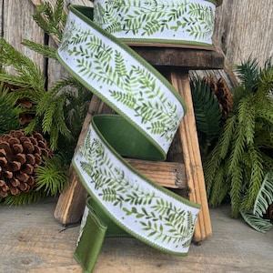 May include: A spool of wired ribbon featuring a white background with a repeating green fern leaf pattern. The ribbon has solid green edges and is coiled in a spiral shape, showcasing the botanical print and the smooth, dark green underside.