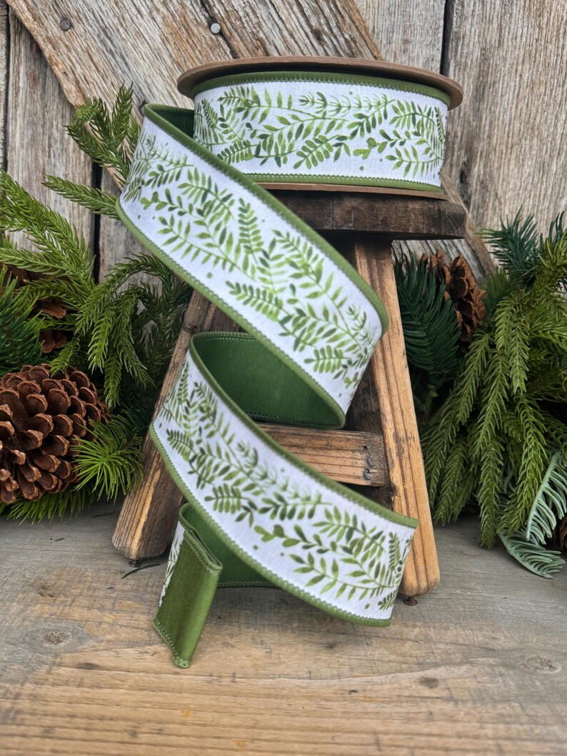 May include: A spool of wired ribbon featuring a white background with a repeating green fern leaf pattern. The ribbon has solid green edges and is coiled in a spiral shape, showcasing the botanical print and the smooth, dark green underside.