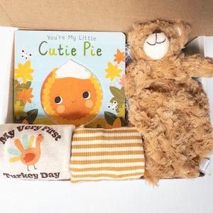 May include: A gift box containing a board book titled You're My Little Cutie Pie with a pie illustration, a tan plush teddy bear, a cream-colored onesie with a turkey graphic and the text My Very First Turkey Day, and striped pants.