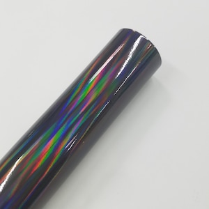 May include: A roll of iridescent vinyl with a dark, glossy finish. The vinyl reflects a spectrum of colors, including green, blue, and purple, creating a holographic effect. The roll is cylindrical and appears to be new.