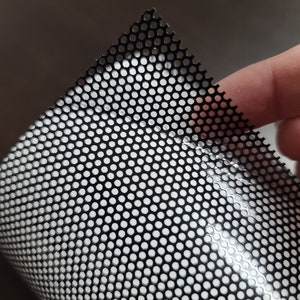 May include: Close-up of a black perforated vinyl sheet with a grid of small, evenly spaced holes. The sheet is partially rolled, revealing a white backing. The texture and pattern suggest a window film or similar product.
