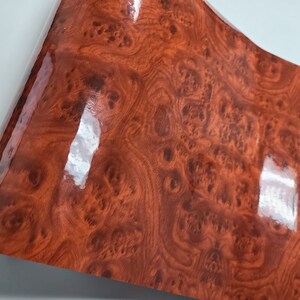 May include: A close-up of a woodgrain vinyl sheet with a glossy finish. The sheet has a reddish-brown color with a swirling pattern.