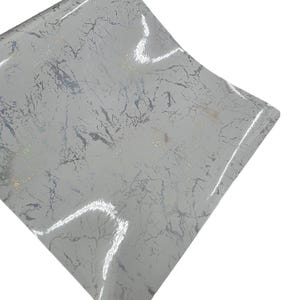 May include: A sheet of light gray vinyl with a marble pattern. The design features silver and gold veining, creating a luxurious and textured appearance. The material has a glossy finish, reflecting light.