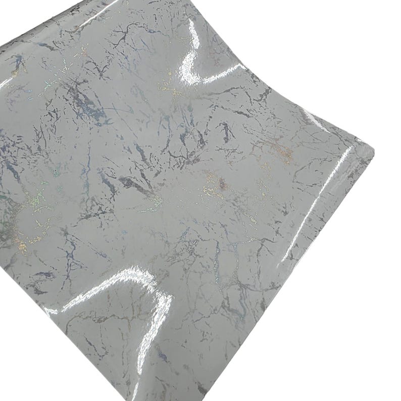 May include: A sheet of light gray vinyl with a marble pattern. The design features silver and gold veining, creating a luxurious and textured appearance. The material has a glossy finish, reflecting light.