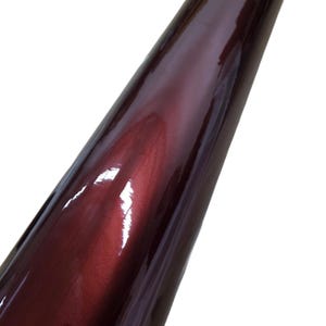 May include: A roll of glossy, burgundy-colored vinyl material. The surface reflects light, creating highlights and shadows. The material appears smooth and is tightly rolled, suggesting it is new and unused. The color is a deep, rich shade of red.
