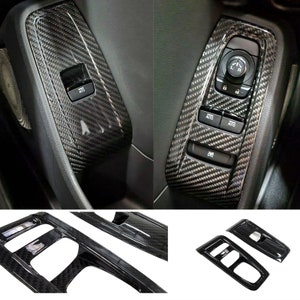 May include: Carbon fiber car interior trim pieces. The image shows several pieces, including window switch panels and console trim, with a glossy, woven carbon fiber pattern. The pieces are designed to fit around existing car components.