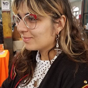 May include: A person with blonde and brown hair wearing a black jacket with red and gold accents. They are wearing a white shirt with black polka dots and a silver necklace with a round pendant. They are also wearing glasses with red accents and a silver earring with a cross and a hand design.