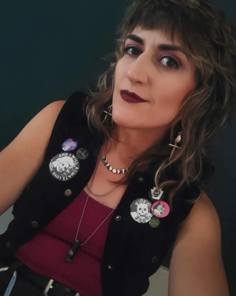 May include: A person wearing a black vest with various pins and patches, including a skull and crossbones pin, a pin with a cat cartoon, and a pin with the text "Video Kill." The vest is over a burgundy tank top. The person is wearing a silver chain necklace with a black stone pendant and a silver chain choker with a moon and star design.