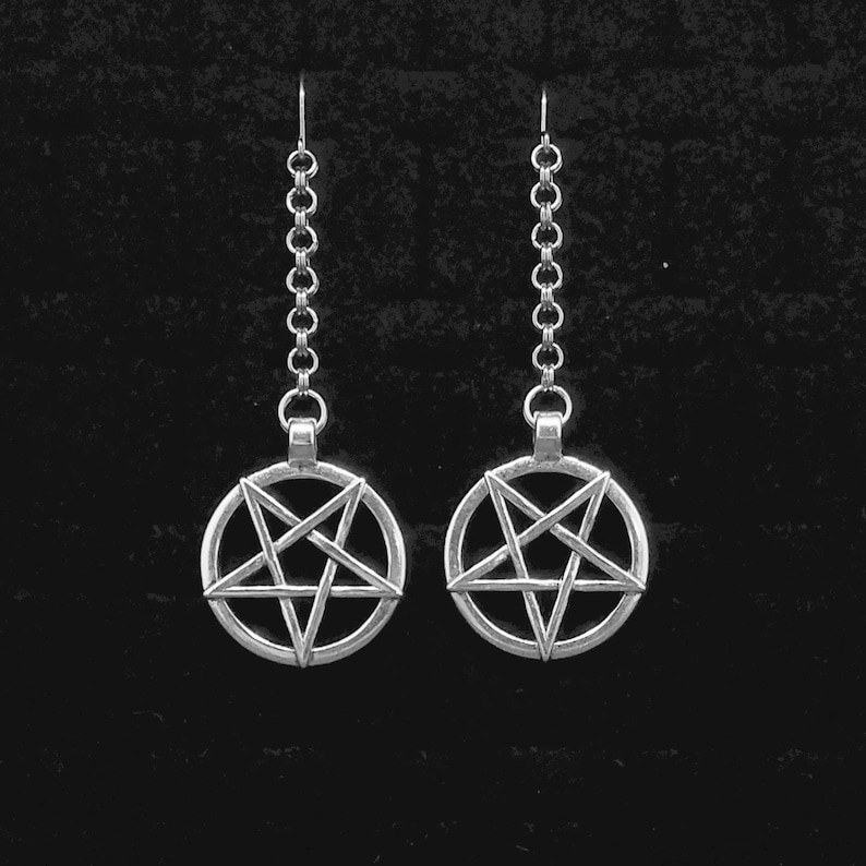 Pu&ograve; includere: Un paio di orecchini d'argento con un ciondolo a pentagramma su ogni orecchino. Gli orecchini sono su uno sfondo nero.