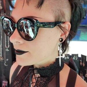 May include: A person with a shaved head and black sunglasses is wearing a black lace top with a black choker and a silver cross earring.
