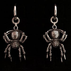 May include: A pair of silver-toned spider earrings. Each earring features a detailed spider pendant with textured body and legs, suspended from a hoop. The earrings are designed with intricate details, showcasing a gothic or alternative aesthetic.
