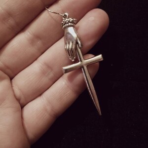 May include: Silver earring featuring a hand holding a dagger that is piercing a cross.