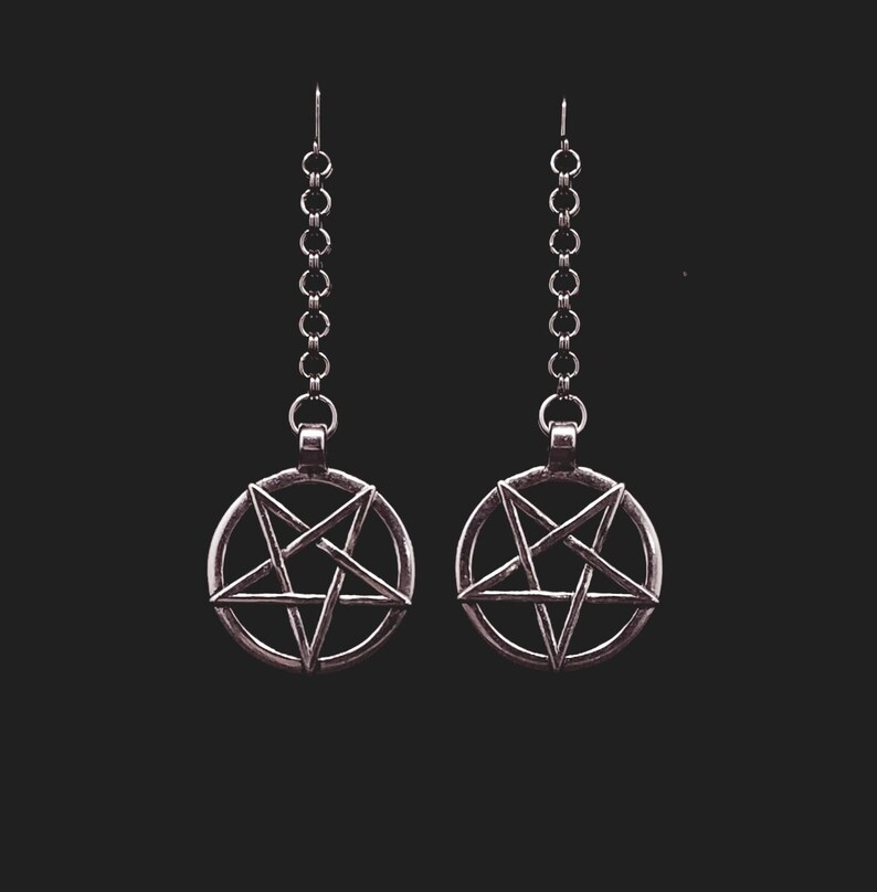 Pu&ograve; includere: Un paio di orecchini d'argento con un ciondolo a pentagramma su ogni orecchino. Gli orecchini sono su uno sfondo nero.