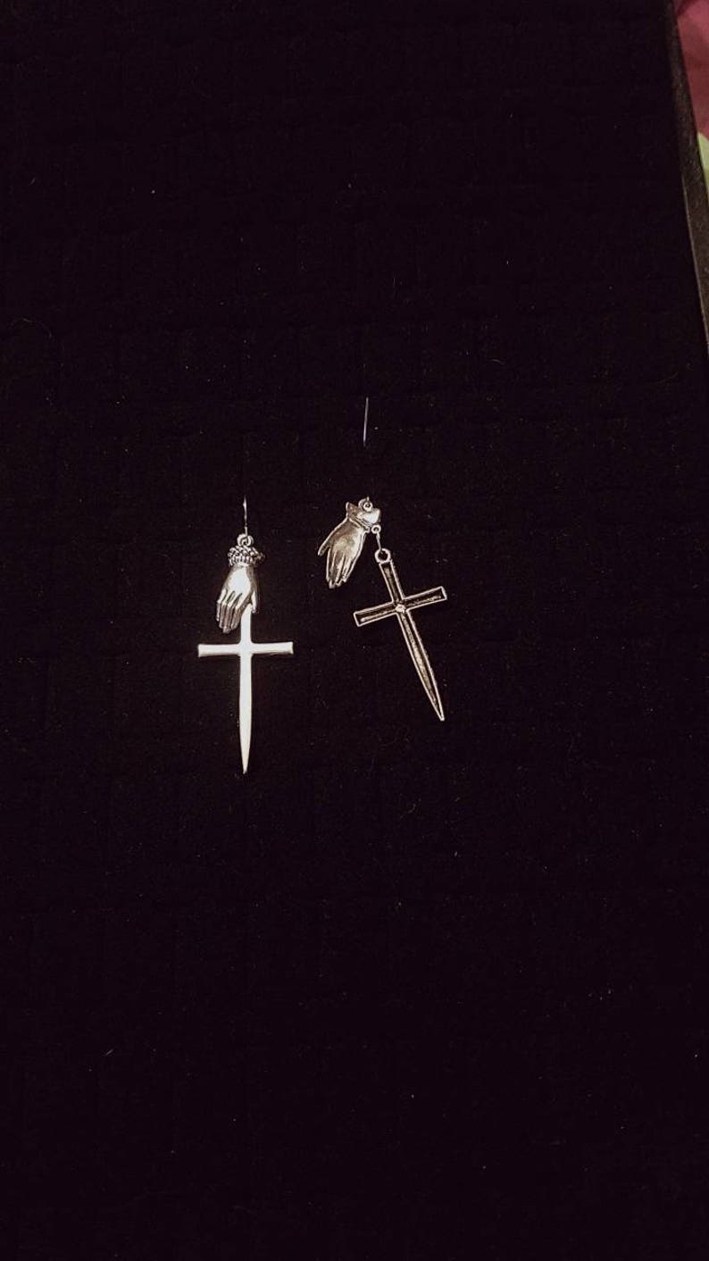 May include: A pair of silver earrings with a cross design. Each earring features a small hand holding the cross.
