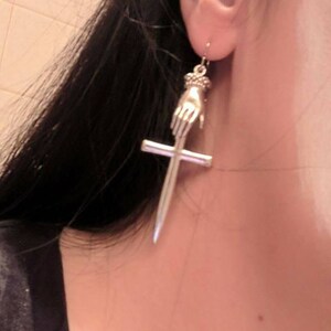 May include: Silver dangle earrings featuring a hand holding a cross. The hand and cross are detailed and have a vintage look.