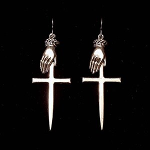 May include: A pair of silver earrings featuring a hand holding a dagger that is positioned on top of a cross.