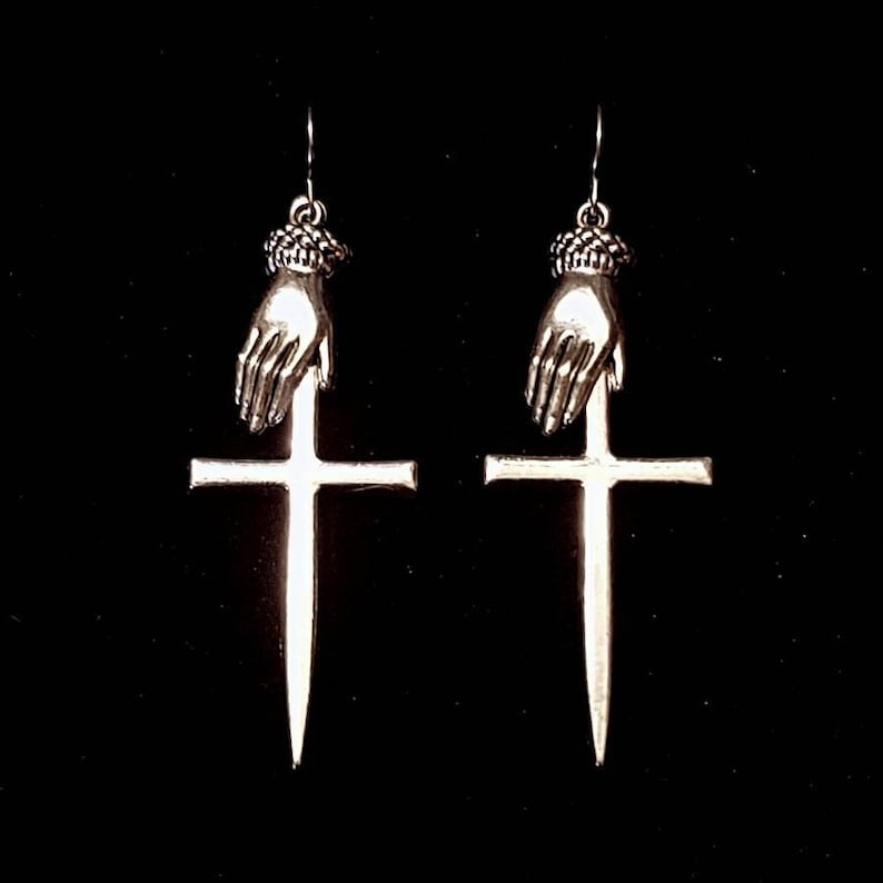 May include: A pair of silver earrings featuring a hand holding a dagger that is positioned on top of a cross.