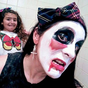 May include: A woman with white face paint and red accents around her eyes and mouth is wearing a black and red plaid headband. She is wearing a silver dagger earring. A young girl with curly brown hair is in the background.