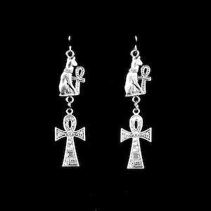 Puede incluir: Pendientes de aro de plata con un gato sosteniendo un símbolo ankh, con un amuleto ankh más grande que cuelga debajo.
