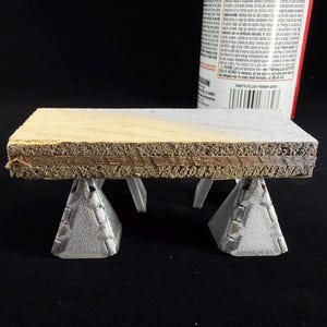 May include: A small, rectangular wooden platform supported by two gray, triangular stands. The platform has a light-colored top and a darker, textured edge. A can of primer spray is partially visible in the background.