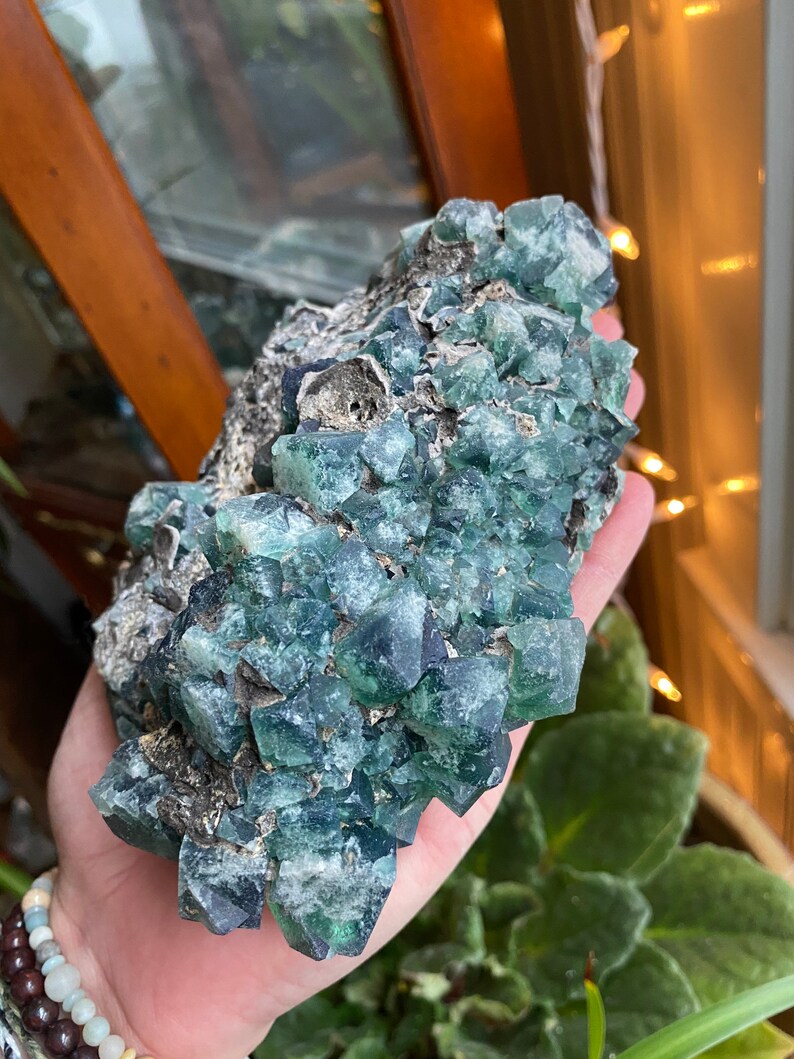May include: A large cluster of green fluorite crystals with a rough, natural texture. The crystals are clustered together in a dense formation, creating a visually interesting and unique piece.