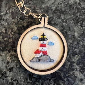 Puede incluir: Un llavero redondo de madera con un diseño de faro bordado. El faro es rojo y blanco con una parte superior negra, sobre rocas grises con nubes azules. El llavero tiene un cierre y un anillo dorados.