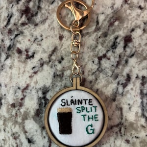 May include: A gold-colored keychain with a round, wooden embroidery hoop pendant. The pendant features the text "Sl&aacute;inte Split the G" and an embroidered image of a dark stout. The keychain has a clasp and a key ring.