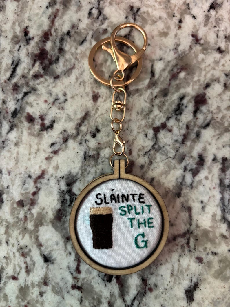 May include: A gold-colored keychain with a round, wooden embroidery hoop pendant. The pendant features the text "Sl&aacute;inte Split the G" and an embroidered image of a dark stout. The keychain has a clasp and a key ring.