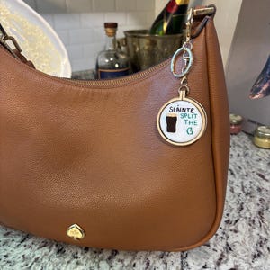May include: A brown leather handbag with a gold-colored keychain. The keychain has a round charm with the words "Sl&aacute;inte Split the G" and a graphic of a pint of stout. The handbag has a gold spade logo.