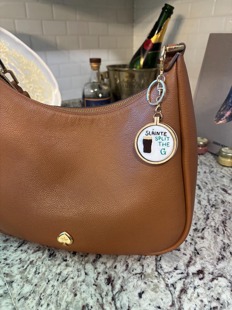 May include: A brown leather handbag with a gold-colored keychain. The keychain has a round charm with the words "Sl&aacute;inte Split the G" and a graphic of a pint of stout. The handbag has a gold spade logo.