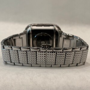 May include: A silver-toned smartwatch with a matching band. The watch face is encrusted with small, sparkling stones. The band features a textured, patterned design, and the watch is set against a plain background.